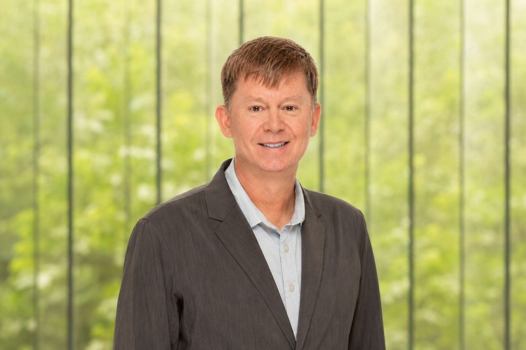 Portrait of a smiling man in a gray suit and light button-down shirt against a blurred green background.